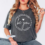 a woman sitting on a chair wearing a t - shirt that says be you